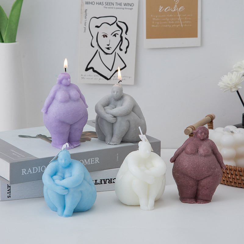 Five body-positive candle figurines in different colors on a surface with books and decor in the background.